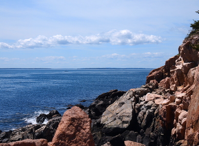 [Multi-colored dark rock on the right with dark ocean waters on the left. Puffy white clouds in an overcast sky above.]